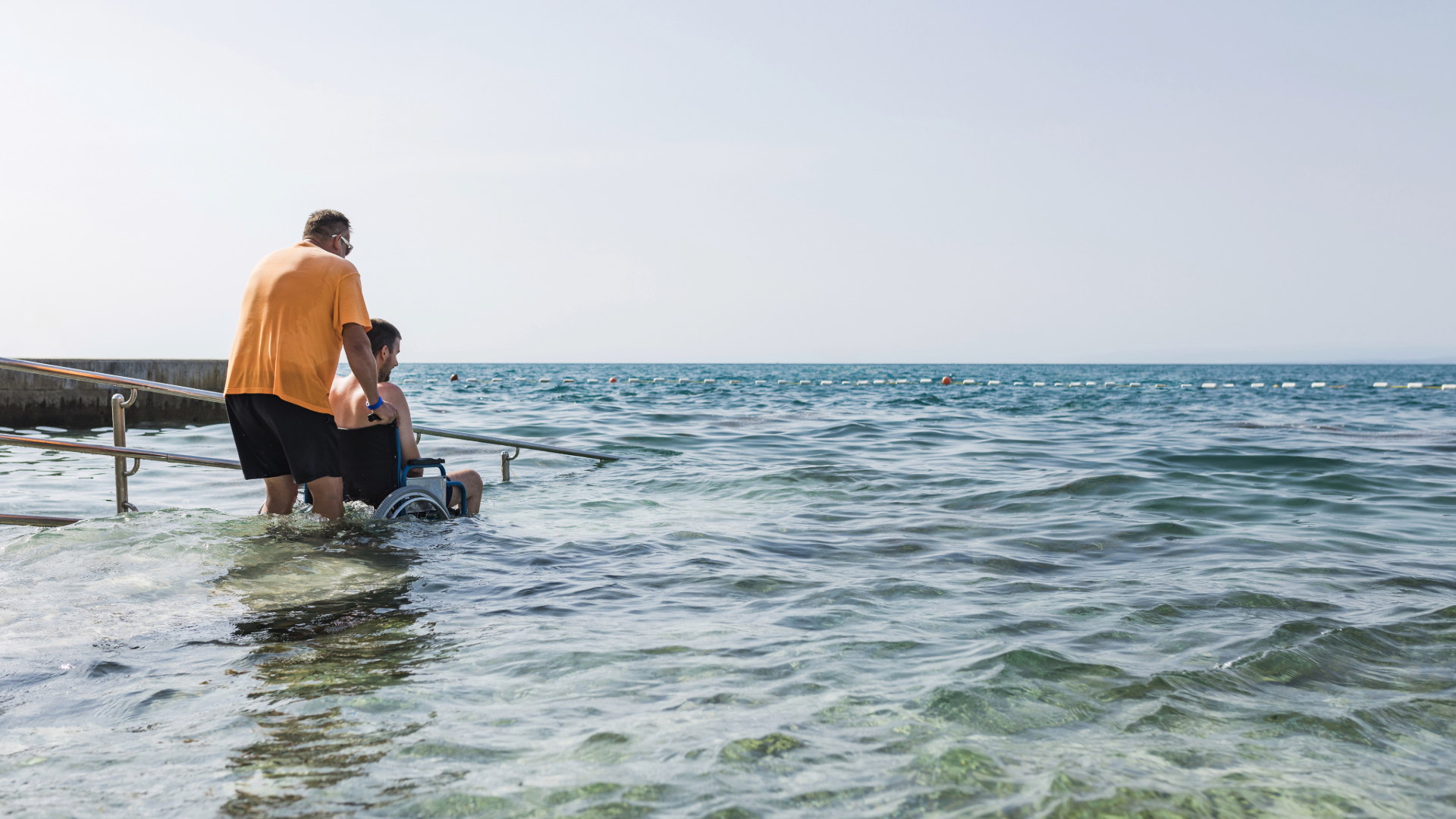 Assistent hjälper person i rullstol ner en ramp på stranden för ett bad.